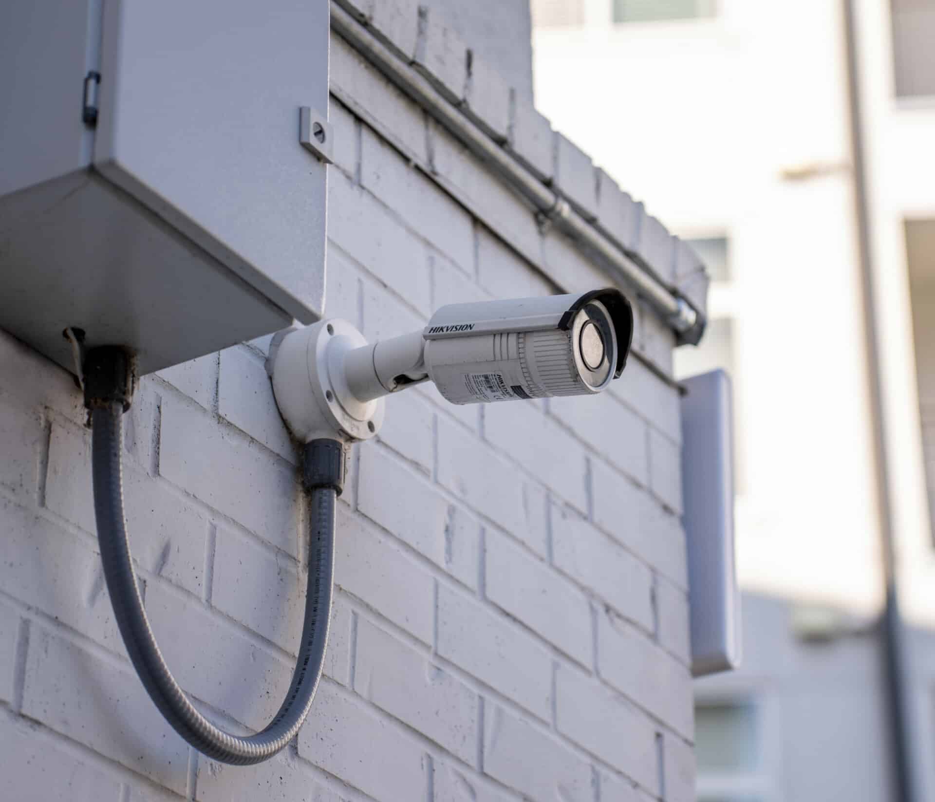 An external security camera is mounted to the control box on the side of a brick building.