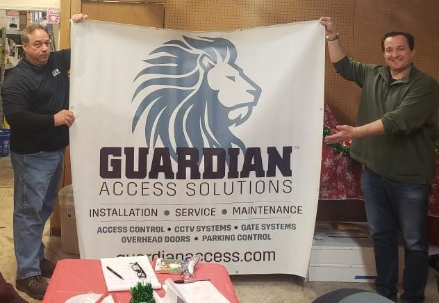 Two people standing on each side of a large canvas sign that reads Guardian Access Solutions.